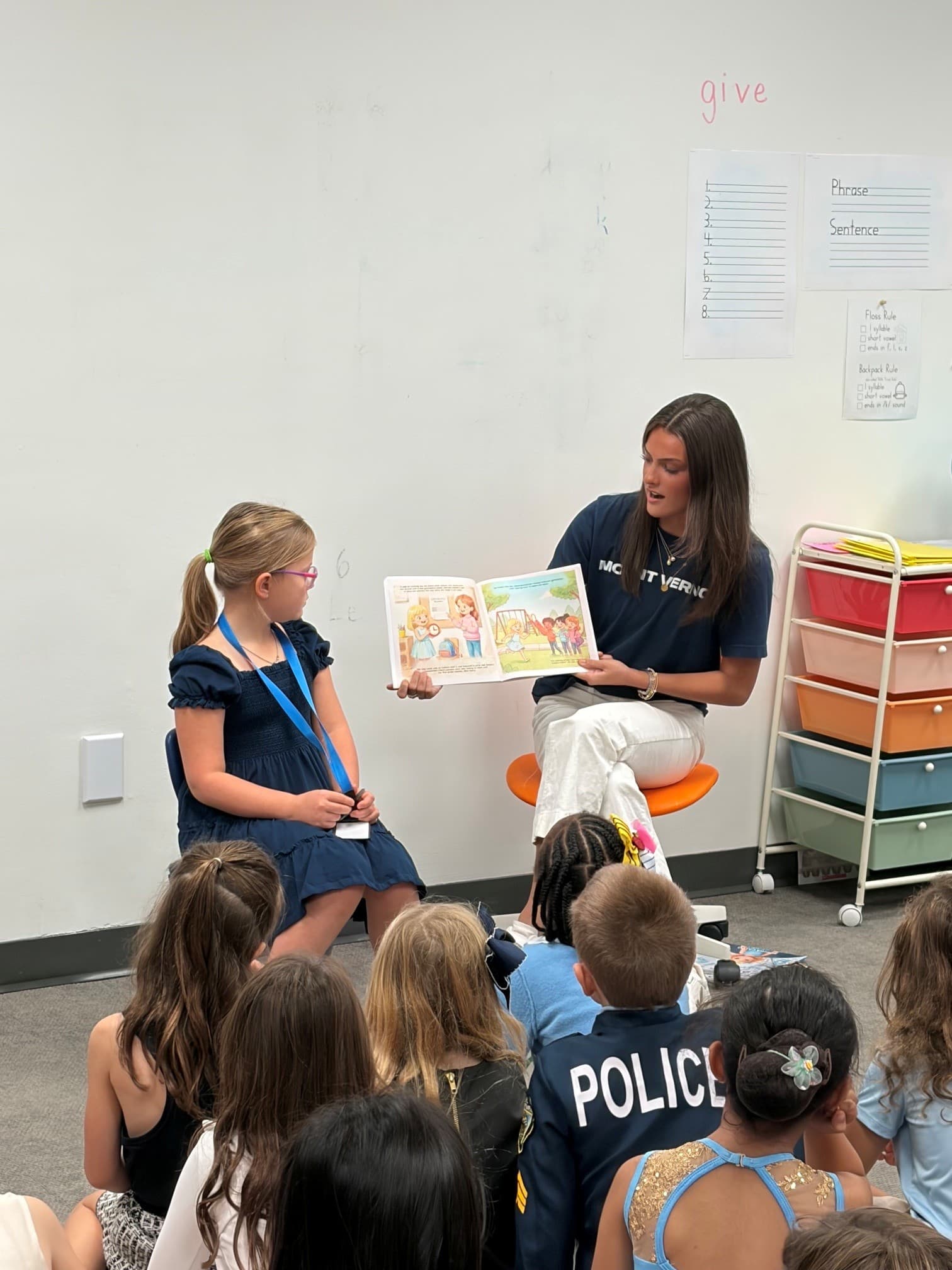A woman reads a picture book to children sitting on the floor in a classroom.