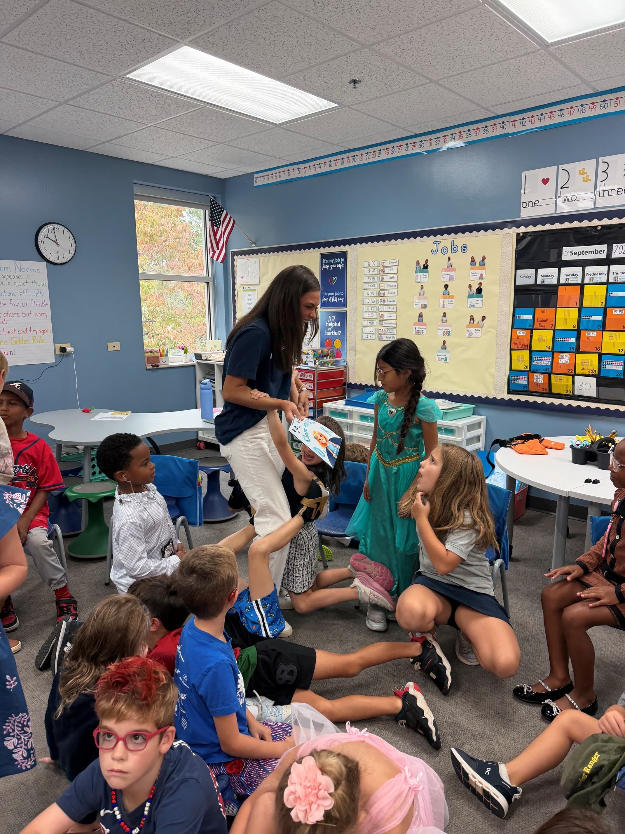 A teacher interacts with diverse young students sitting on a colorful classroom floor.