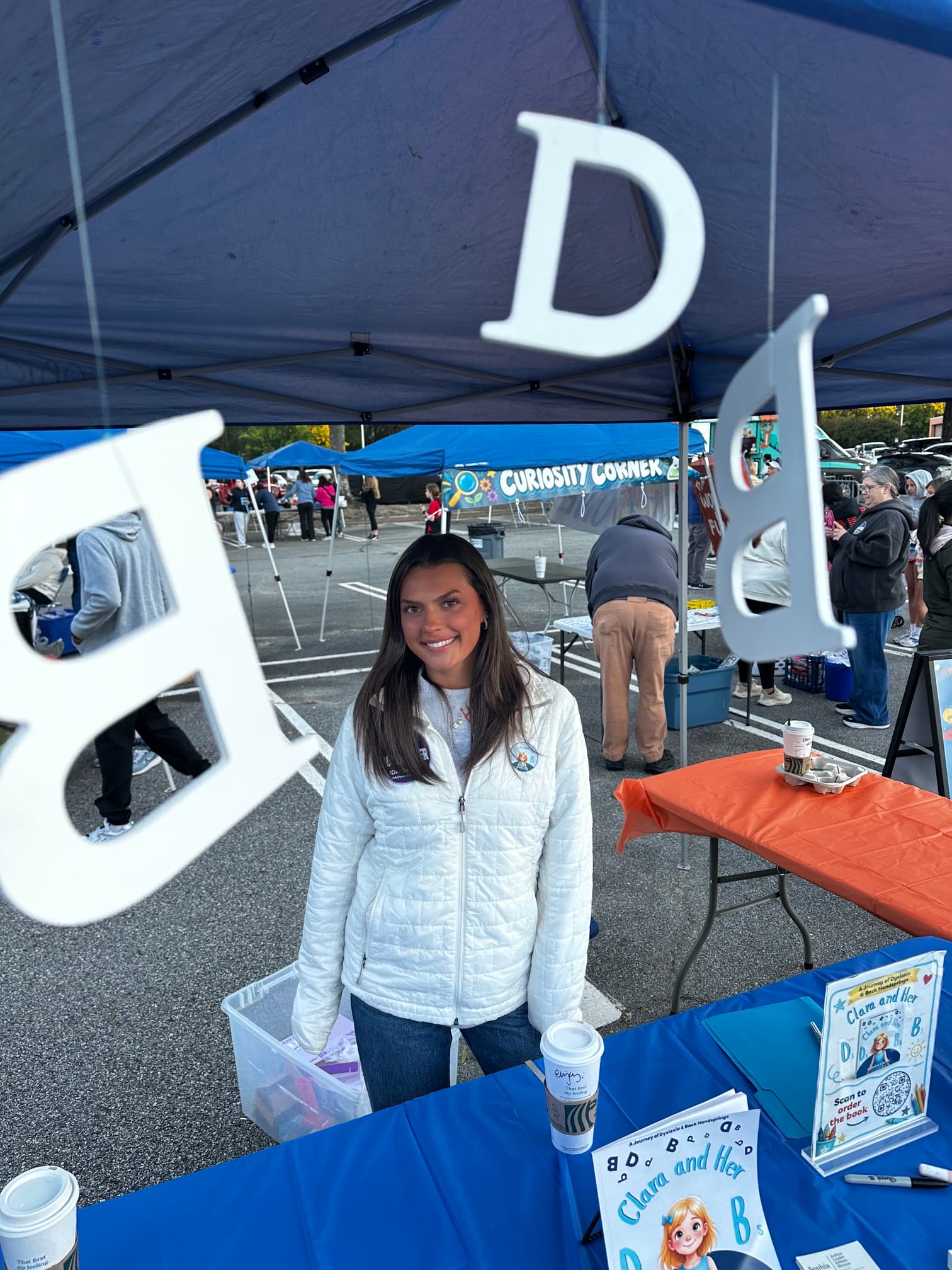 Smiling woman in a white jacket behind a book table with large hanging letters.